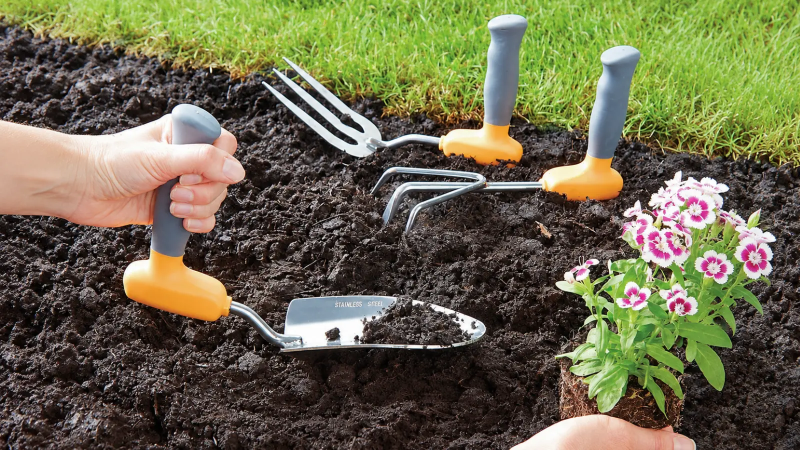 Hand using an ergonomic garden trowel with a vertical grip beside similarly designed hand tools and a flowering plant ready to be planted in prepared garden soil