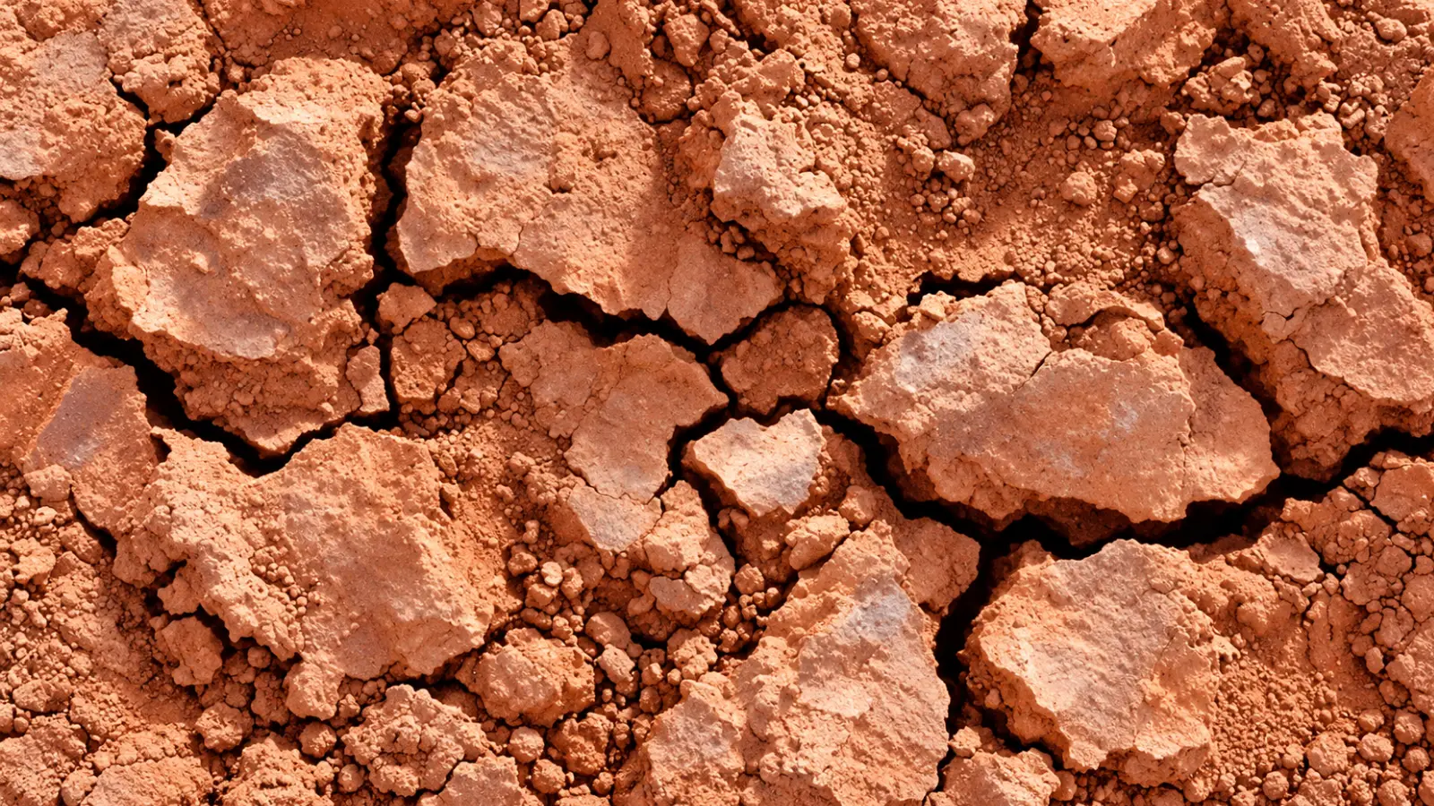 Close-up of dry reddish clay soil with large hardened clumps, smaller compacted particles and rough cracked surfaces, showing the dense texture of clay-rich ground